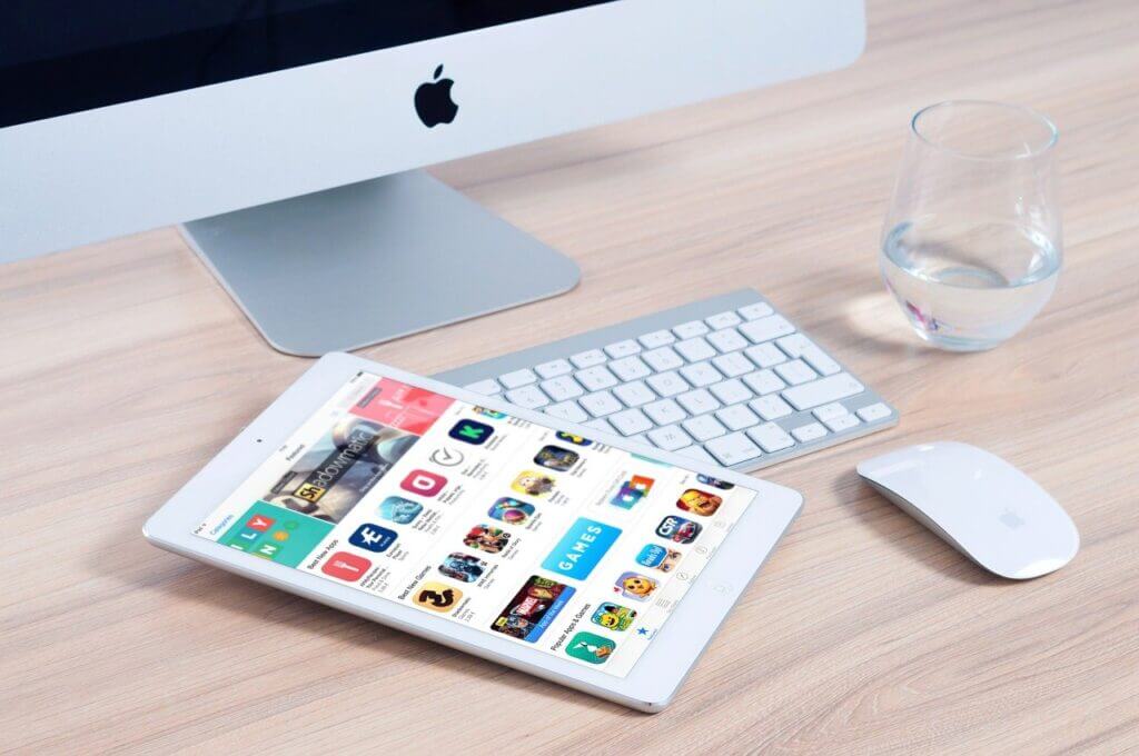 iMac with a keyboard, mouse and white iPad on a desk beside a glass of water.
