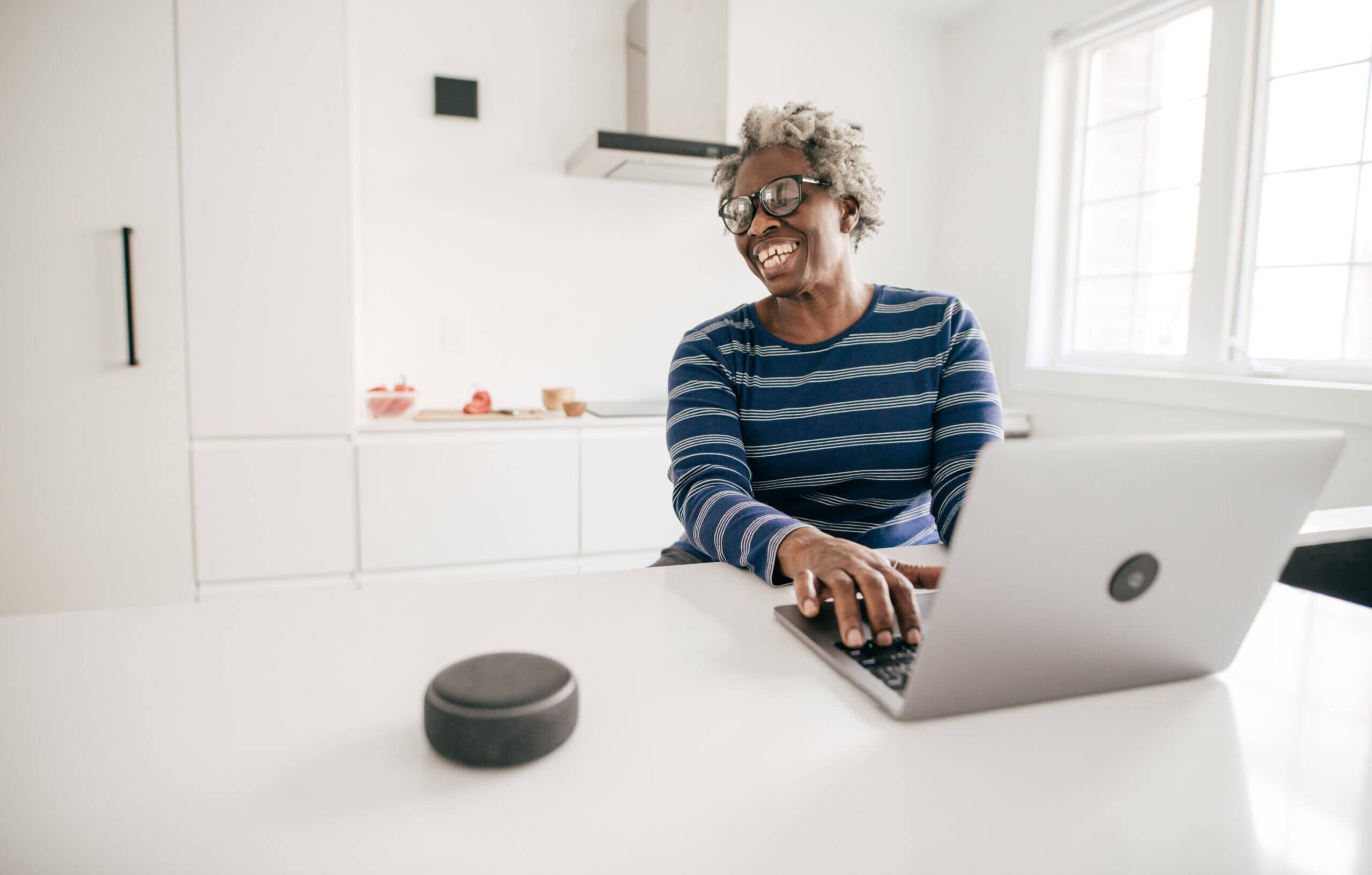 Woman smiling while sitting at kitchen counter with laptop and smart speaker.
