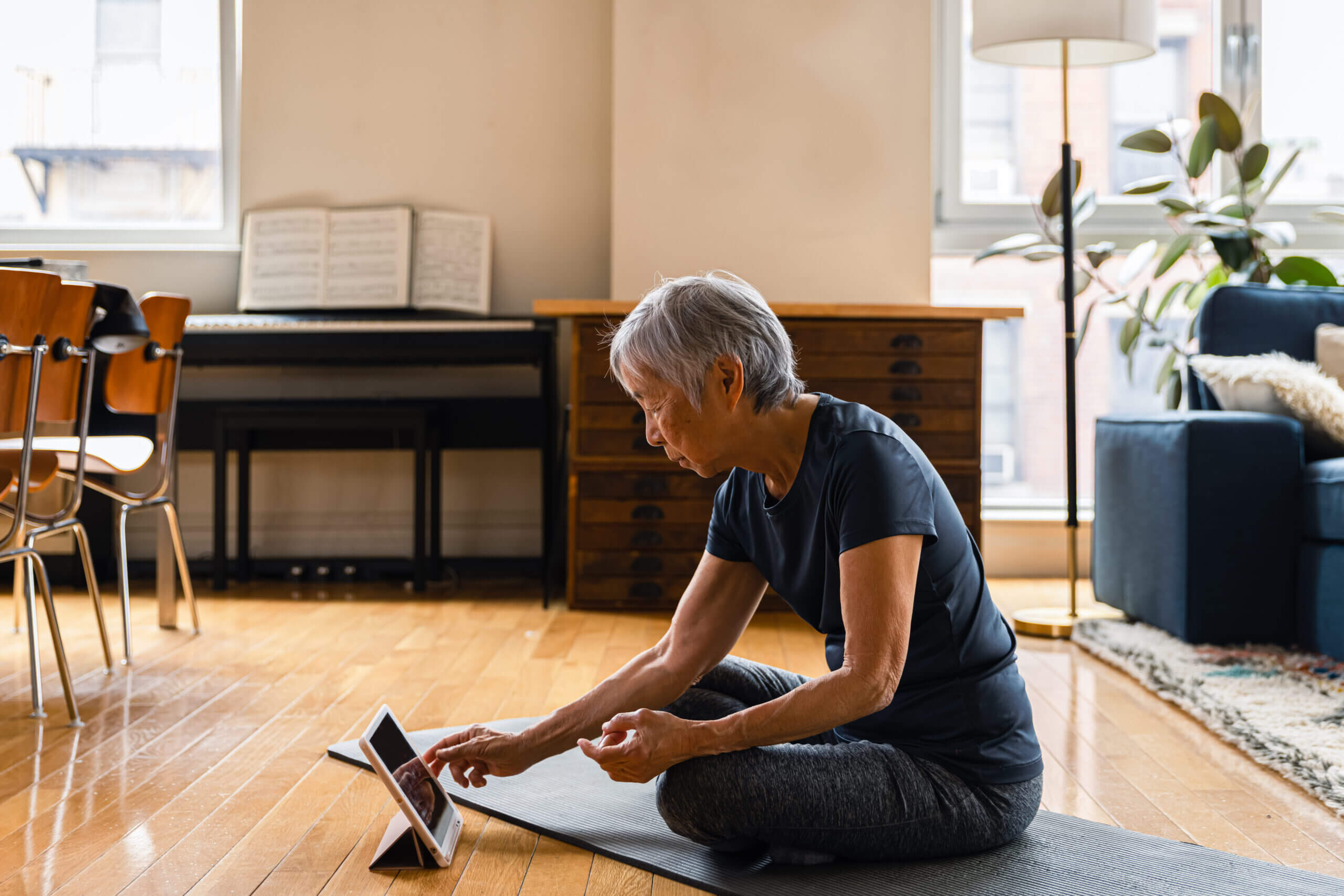 Older woman sitting on yoga mat looking at a tablet.